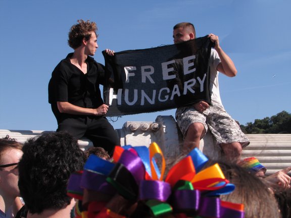 Zwei Personen halten eine „FREE HUNGARY“-Flagge bei der Budapest Pride 2025 hoch, während sie auf dem Brückenpfeiler der Elisabethbrücke sitzen. © Creative Commons Attribution 4.0 International